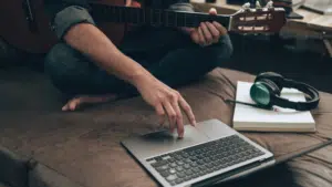 A person is comfortably sitting on a sofa, laptop balanced nearby and a guitar beside them, with headphones resting around their neck, ready to dive into the world of SplitShare for seamless creative sharing.