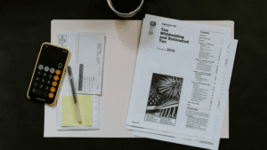 Amidst tax documents and forms for 2019, a smartphone with a calculator app open sits on the desk beside a pen, an envelope, and a cup of coffee. As an independent musician juggling royalties and expenses, preparing taxes requires focused attention.