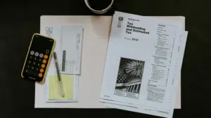 Amidst tax documents and forms for 2019, a smartphone with a calculator app open sits on the desk beside a pen, an envelope, and a cup of coffee. As an independent musician juggling royalties and expenses, preparing taxes requires focused attention.