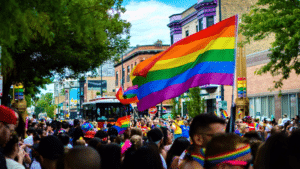 A crowd of prideful people walking down a street with a rainbow flag.