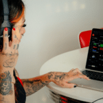 A person with tattoos, deeply engrossed in their work, wears headphones and uses a laptop at a table, mastering the art of social media marketing.