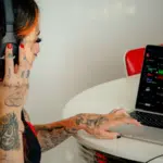 A person with tattoos, deeply engrossed in their work, wears headphones and uses a laptop at a table, mastering the art of social media marketing.