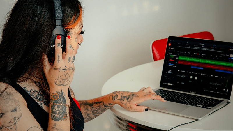 A person with tattoos, deeply engrossed in their work, wears headphones and uses a laptop at a table, mastering the art of social media marketing.