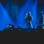 Two musicians perform on stage under blue lighting, their captivating tunes filling the air while a sea of phones rises, creating a wave of UGC as fans eagerly capture the moment.