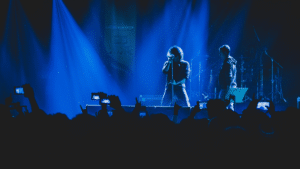 Two musicians perform on stage under blue lighting, their captivating tunes filling the air while a sea of phones rises, creating a wave of UGC as fans eagerly capture the moment.