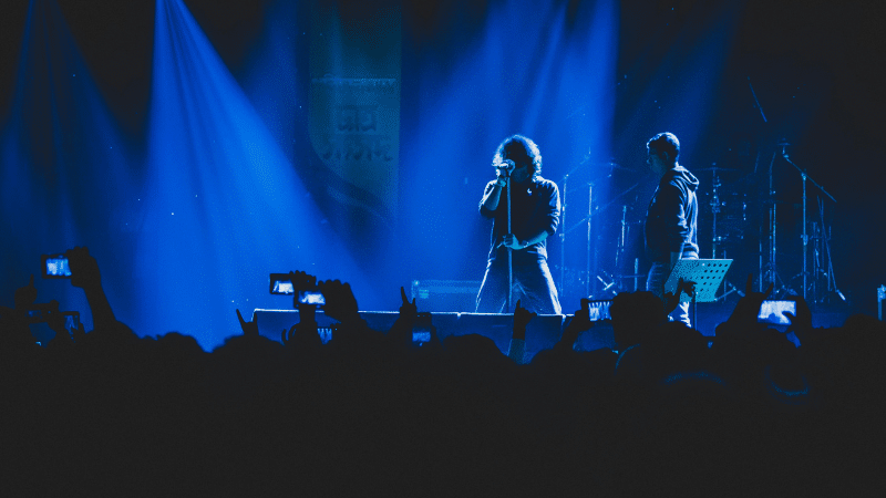 Two musicians perform on stage under blue lighting, their captivating tunes filling the air while a sea of phones rises, creating a wave of UGC as fans eagerly capture the moment.