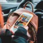 A woman in a brown backpack holding a cell phone in her car, engaged in social media.