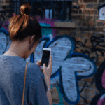 A woman scrolling through her phone in front of a wall covered in graffiti.