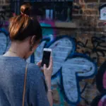 A woman scrolling through her phone in front of a wall covered in graffiti.