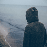 A melancholic man in a hoodie staring at the vast ocean, reflecting his winter blues.