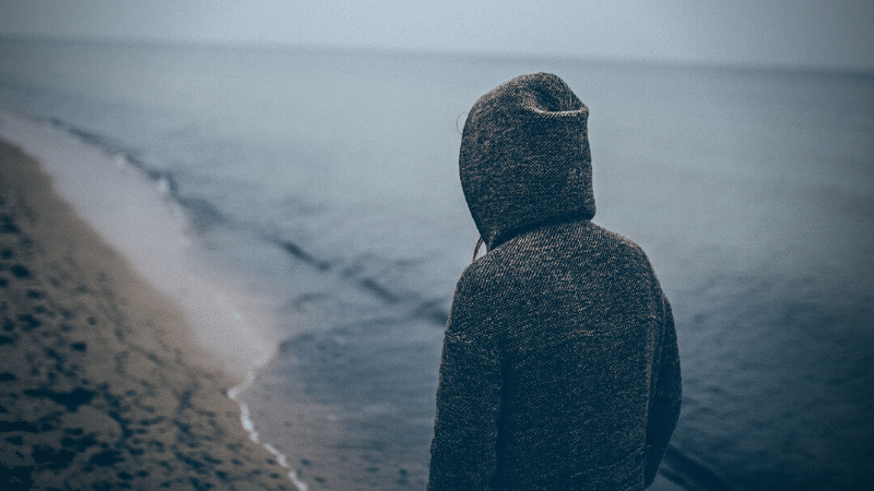 A melancholic man in a hoodie staring at the vast ocean, reflecting his winter blues.