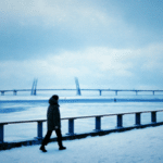A person in winter clothing walks along a snowy path beside a frozen body of water, the large bridge in the background framed by a cloudy sky hinting at winter blues.