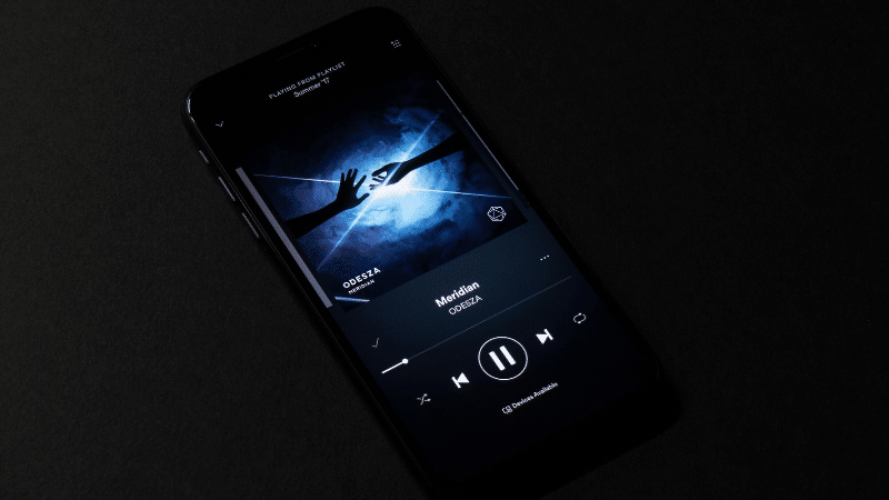 A smartphone screen displays a music player app with a dark-themed interface, featuring the song "Meridian" by ODESZA. The album art shows a hand reaching towards a blue light, perfectly framed on the label page.