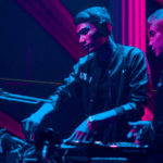 Two starter DJs playing music in front of a neon light.