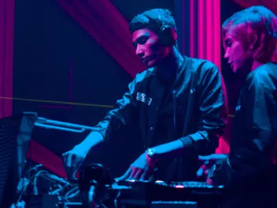 Two starter DJs playing music in front of a neon light.