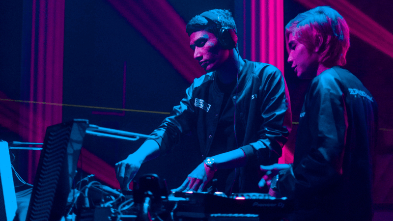 Two starter DJs playing music in front of a neon light.