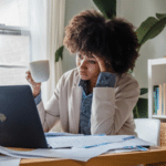 A burnt-out woman sitting at a desk with a laptop and a cup of coffee.