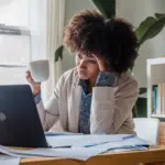 A burnt-out woman sitting at a desk with a laptop and a cup of coffee.