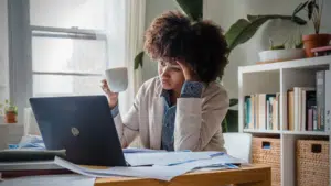 A burnt-out woman sitting at a desk with a laptop and a cup of coffee.