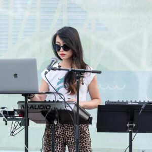 Woman performing women-led music with a laptop and a keyboard at an outdoor event.
