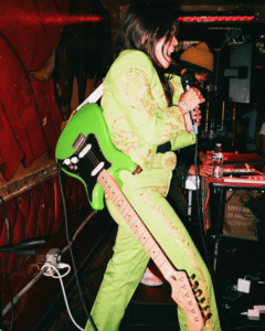 Guitarist in green embroidered suit performing enthusiastically onstage in a women-led music ensemble.