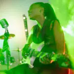 A woman singer playing a guitar in front of green smoke.