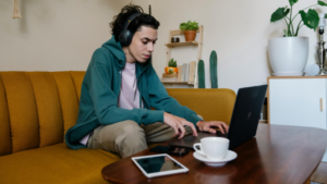 Person wearing headphones sits on a yellow sofa, using a laptop to focus on playlist pitching, with a tablet and a cup nearby. Plants and shelves decorate the background.