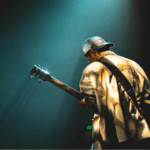 A lone guitarist in a spotlight plays on stage, viewed from behind against a dark, moody background representing the struggles of mental health, with glowing effects.