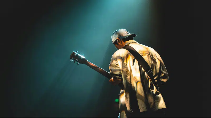 A lone guitarist in a spotlight plays on stage, viewed from behind against a dark, moody background representing the struggles of mental health, with glowing effects.