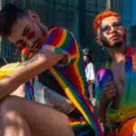 Two people wearing rainbow-themed LGBTQ clothing and accessories sit outdoors at an event, with others and tall buildings visible in the background.