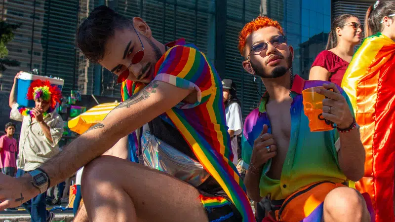 Two people wearing rainbow-themed LGBTQ clothing and accessories sit outdoors at an event, with others and tall buildings visible in the background.