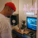 In a music studio, a person in a red beanie and white shirt is deeply immersed in their work, adjusting audio equipment with precision. Nearby, a computer and monitor hint at creative possibilities, perhaps even exploring Spotify Marquee strategies for promoting new sounds.