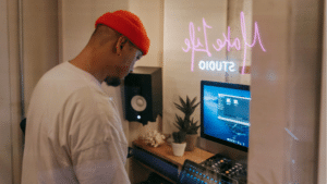 In a music studio, a person in a red beanie and white shirt is deeply immersed in their work, adjusting audio equipment with precision. Nearby, a computer and monitor hint at creative possibilities, perhaps even exploring Spotify Marquee strategies for promoting new sounds.