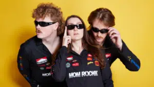 Three individuals in sunglasses showcasing fresh new music on a yellow background.