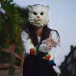 A girl, wearing a cat mask, sits on a fence while enjoying fresh new music.