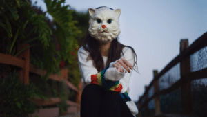 A girl, wearing a cat mask, sits on a fence while enjoying fresh new music.