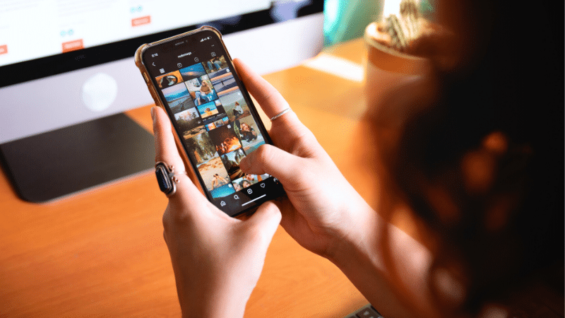 A woman using Instagram on her smartphone while sitting in front of a computer.