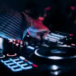A person operates a DJ mixer and turntable with illuminated controls in a dimly lit environment at the Amsterdam Dance Event.