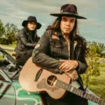 Two people wearing hats and dark clothing pose with a guitar on the hood of a graffiti-covered car in a grassy outdoor area, capturing the vibe of an indie Spotify album cover.