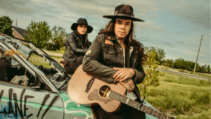 Two people wearing hats and dark clothing pose with a guitar on the hood of a graffiti-covered car in a grassy outdoor area, capturing the vibe of an indie Spotify album cover.