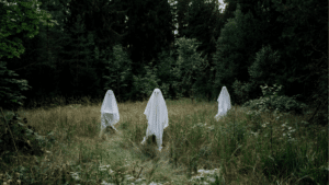 Three people dressed as ghosts with white sheets stand in a grassy, wooded area surrounded by trees—perfect inspiration for your next spooky szn playlist.