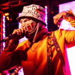 A performer in a gold mask and orange jacket singing into a microphone on a brightly lit stage to promote their new album.