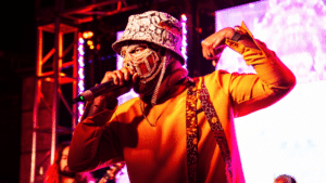 A performer in a gold mask and orange jacket singing into a microphone on a brightly lit stage to promote their new album.