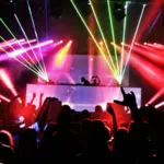 A crowd faces a DJ booth at a concert, as colorful laser lights and smoke illuminate the dark venue, capturing the vibrant energy of New York music.