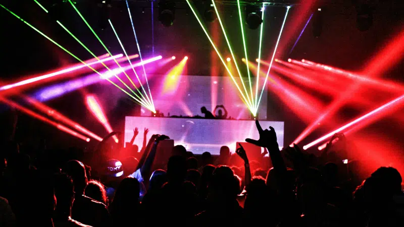 A crowd faces a DJ booth at a concert, as colorful laser lights and smoke illuminate the dark venue, capturing the vibrant energy of New York music.