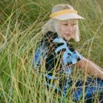 A girl sitting in tall grass wearing a hat while listening to electronic music.