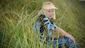 A girl sitting in tall grass wearing a hat while listening to electronic music.