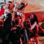 A group of people laughing and raising drinks in a retro-styled room, filled with festive decorations. They're gathered around a couch, reminiscing about holiday songs you can cover for free as they celebrate together.