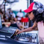 Person wearing headphones and a red cap is DJing at an outdoor event with a laptop and mixer, while a crowd gathers in the background, showcasing perfect gift ideas for musicians.