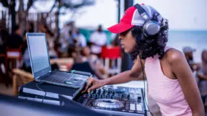 Person wearing headphones and a red cap is DJing at an outdoor event with a laptop and mixer, while a crowd gathers in the background, showcasing perfect gift ideas for musicians.